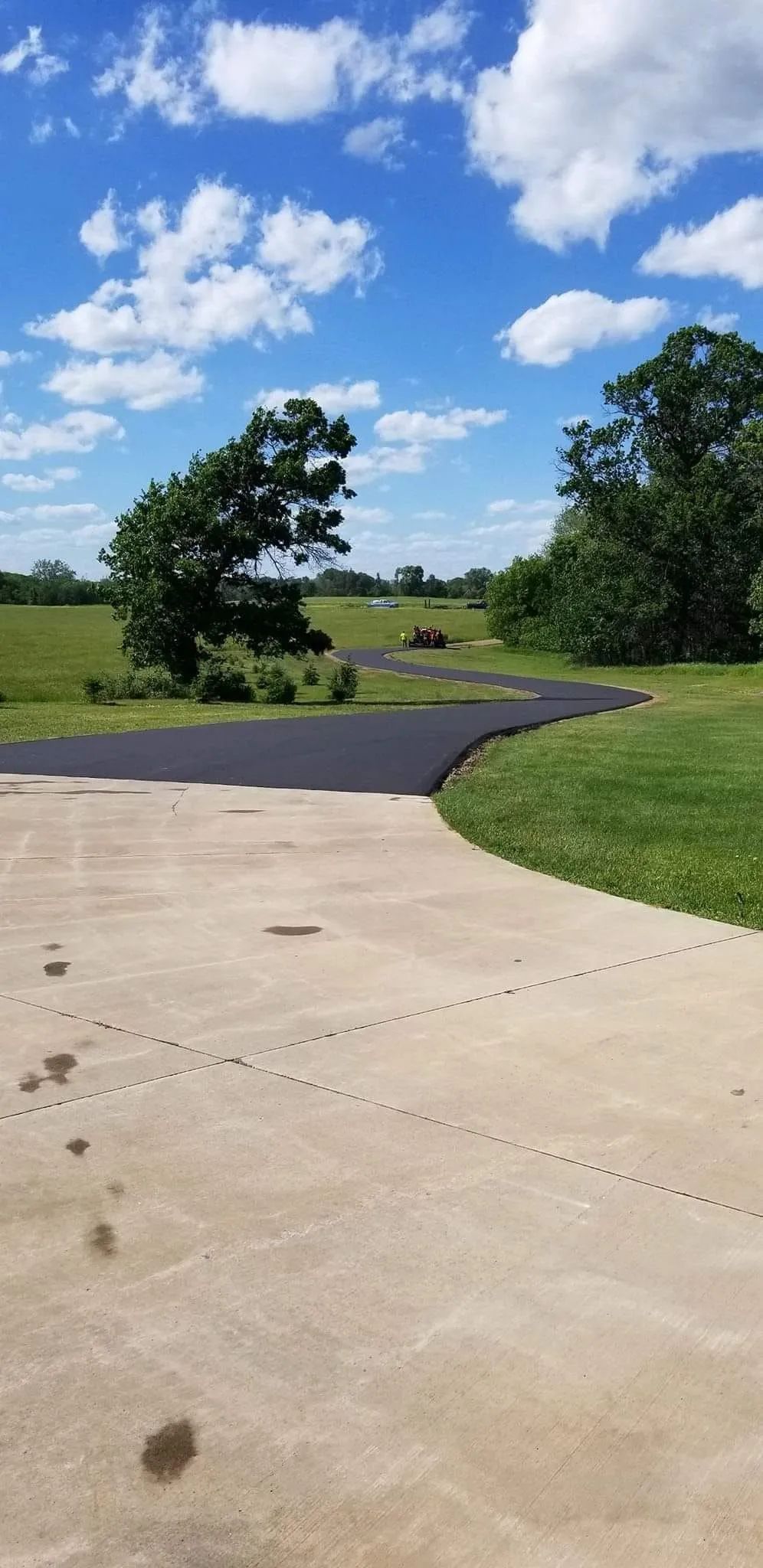 A winding black asphalt driveway transitions into a concrete pathway under a blue sky with clouds.
