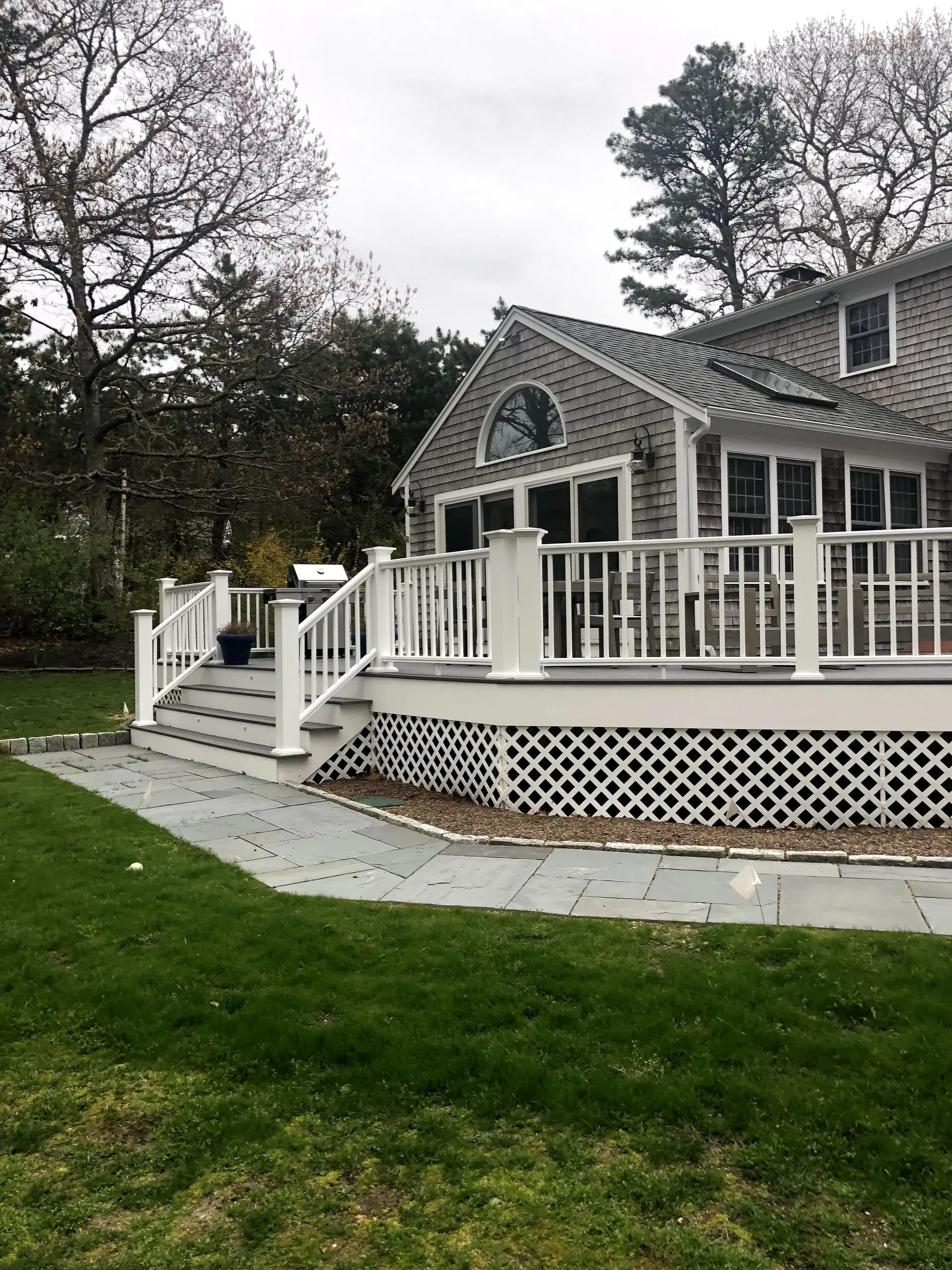 A white-railed deck with stairs attached to a gray-shingled house, set on green grass.