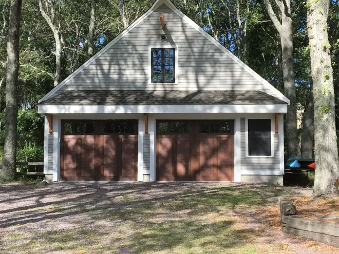 Two-car garage with brown doors, a small window, and a gabled roof, set amongst trees.