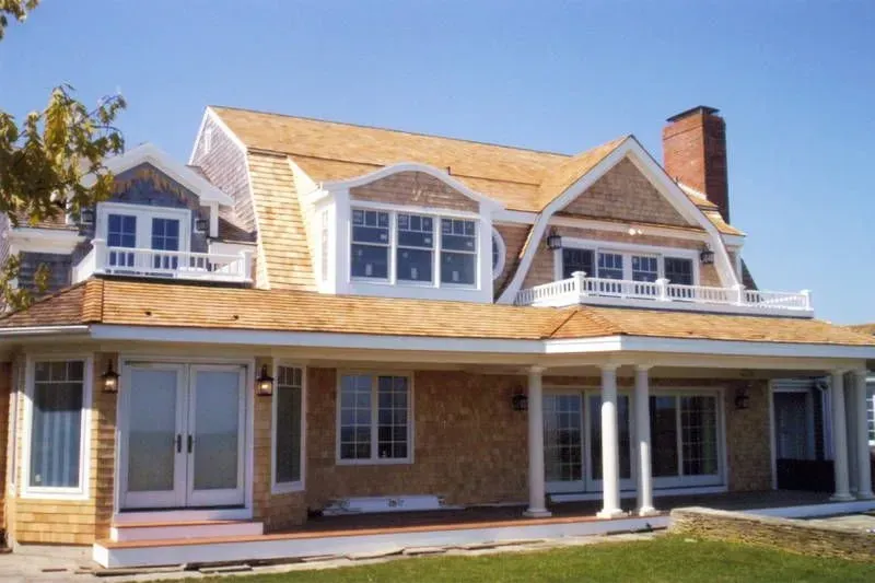 Two-story house with cedar shake siding, dormers, and a wraparound porch; blue sky.