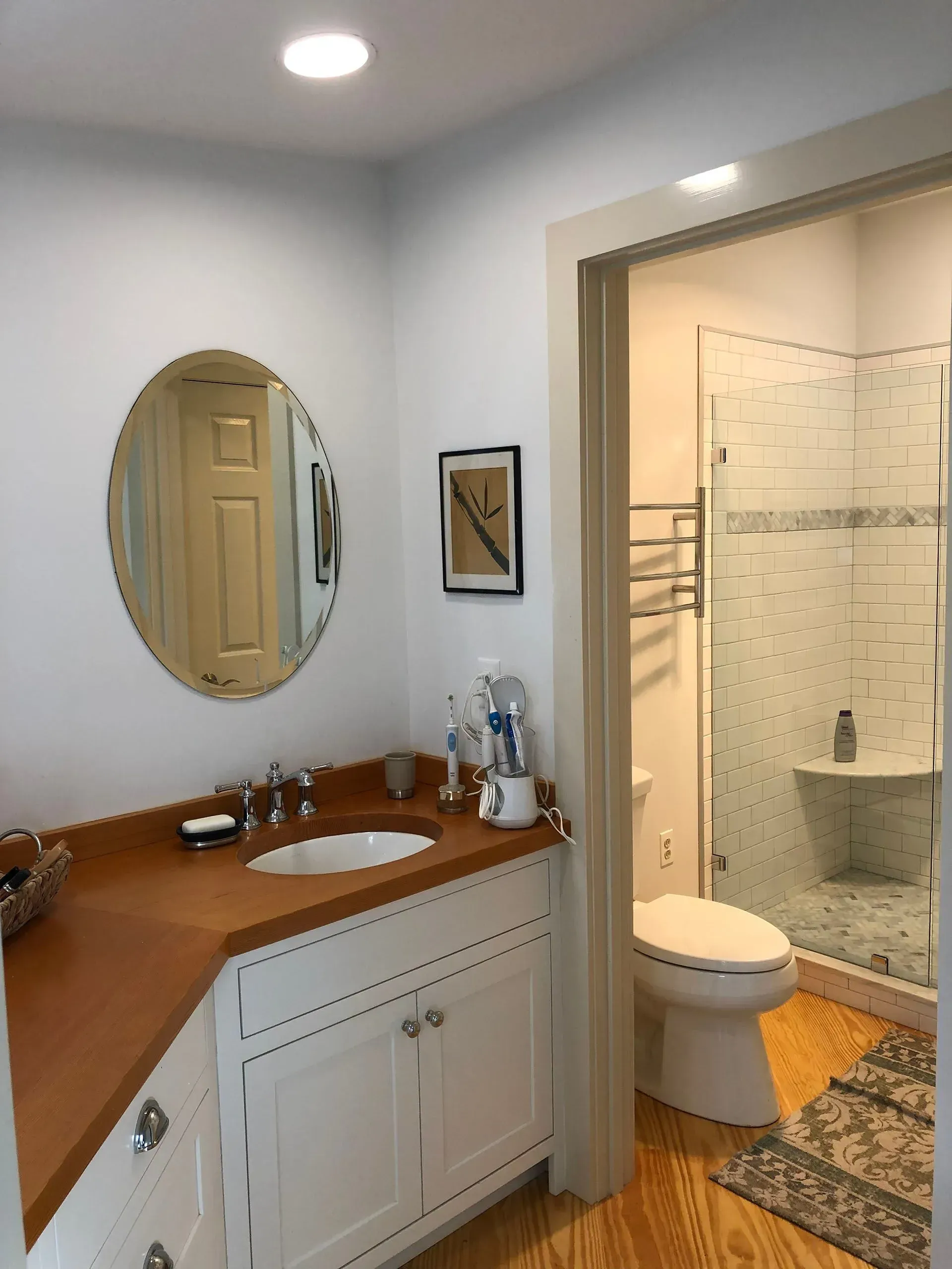Bathroom with white cabinets, wood countertop, oval mirror, shower, and toilet.