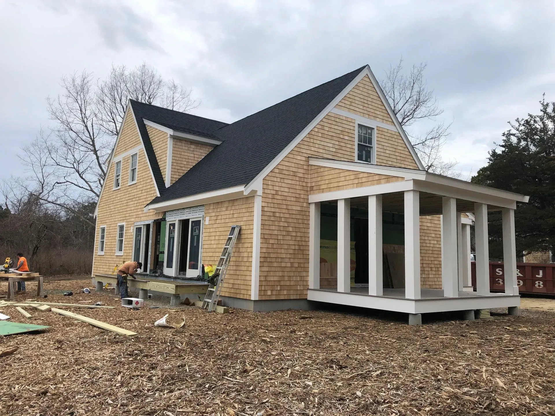 House under construction, cedar shingle siding, dark roof, porch with white columns, cloudy day.