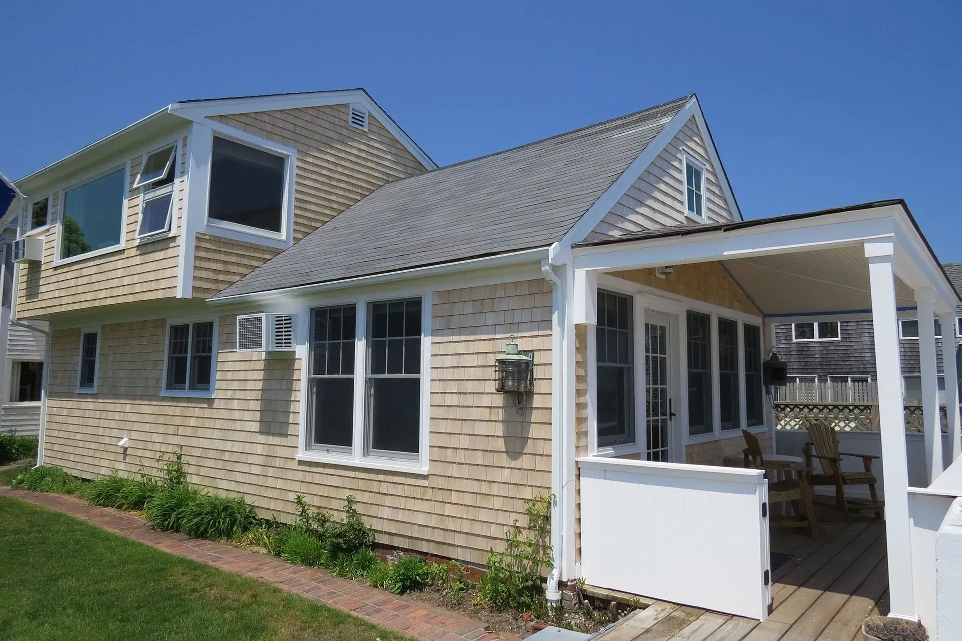 Tan shingled cottage with white trim, porch, and gray roof under a clear blue sky.