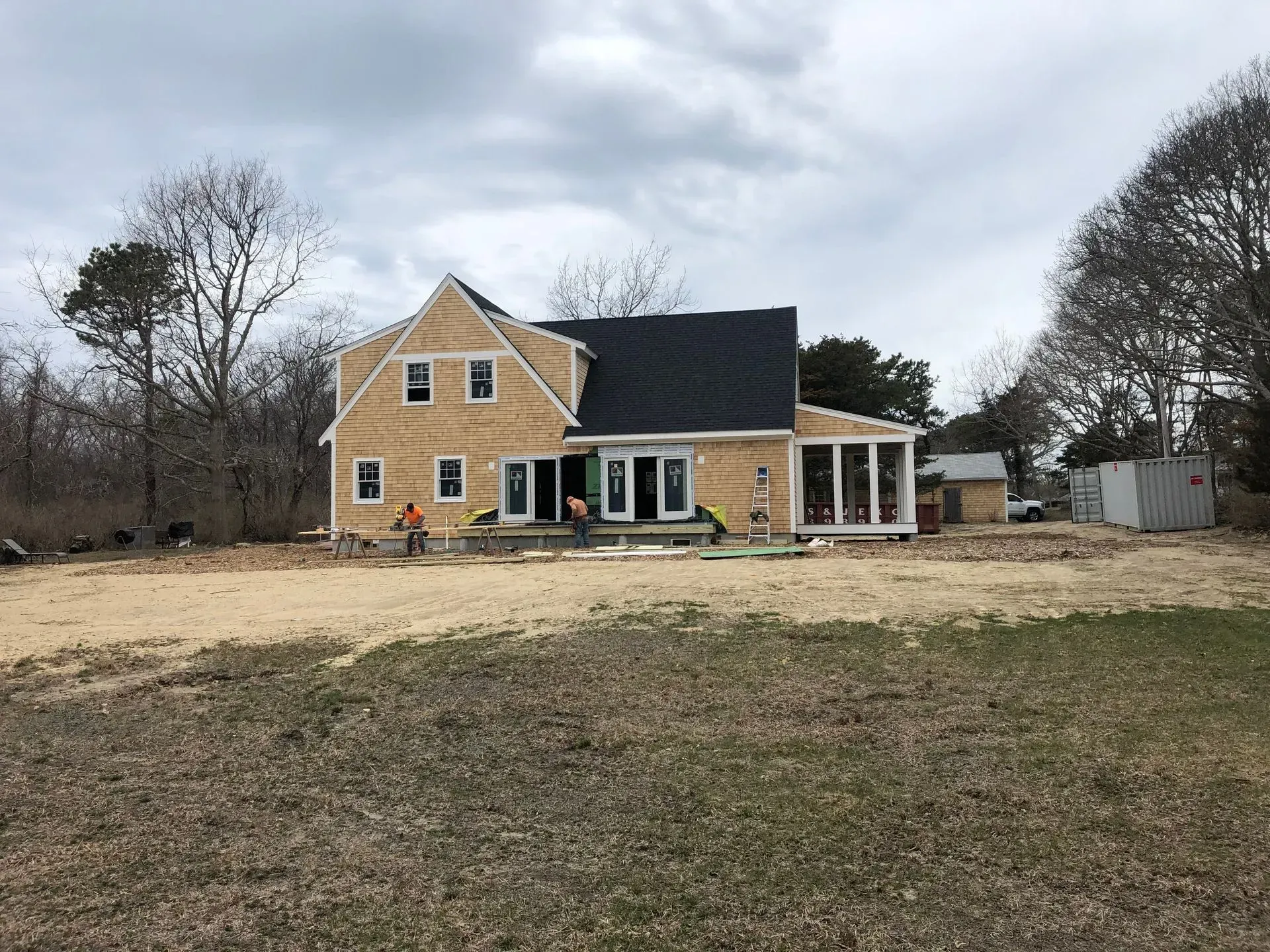 House under construction, yellow siding, black roof, surrounded by trees and a brown field.