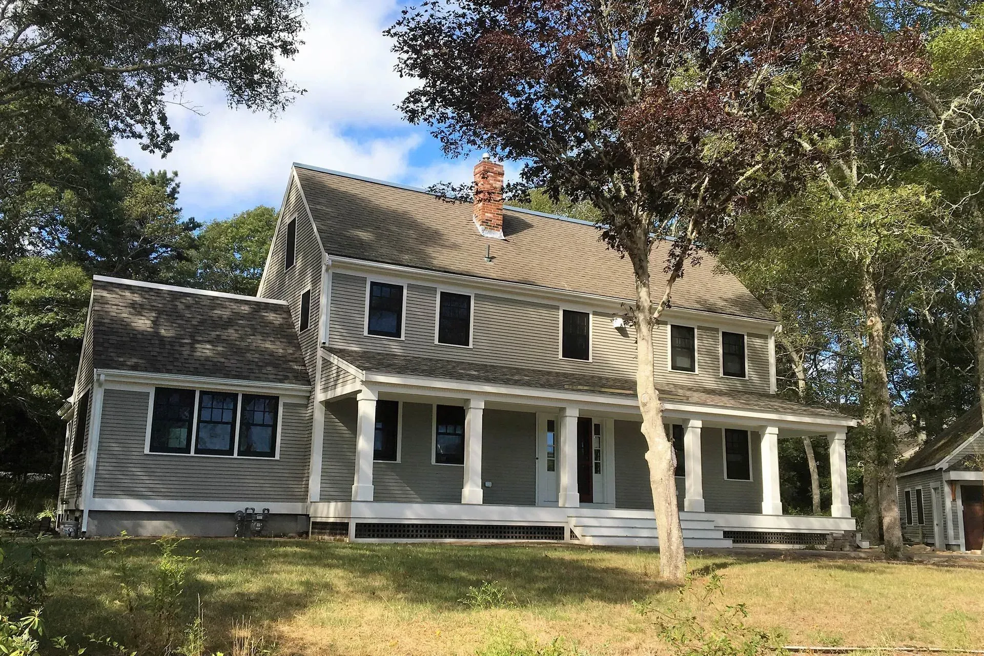 Two-story gray house with white porch, columns, and a chimney, surrounded by trees.