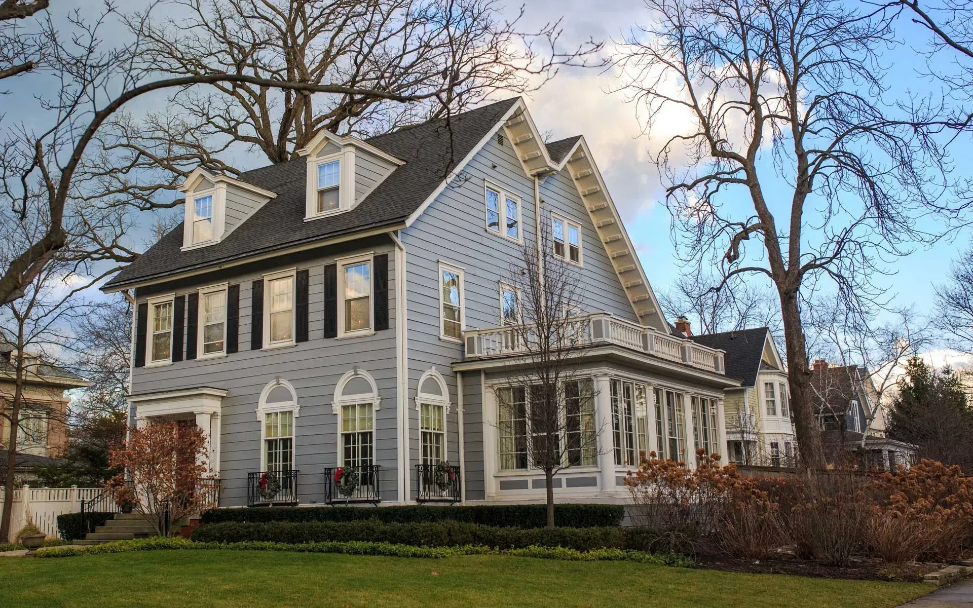 Two-story blue house with black shutters, a porch, and dormers under a bare tree.