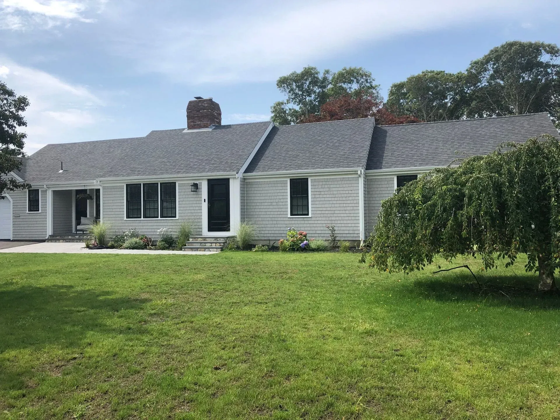 Single-story house with gray siding, dark roof, chimney, green lawn, and a small weeping tree.