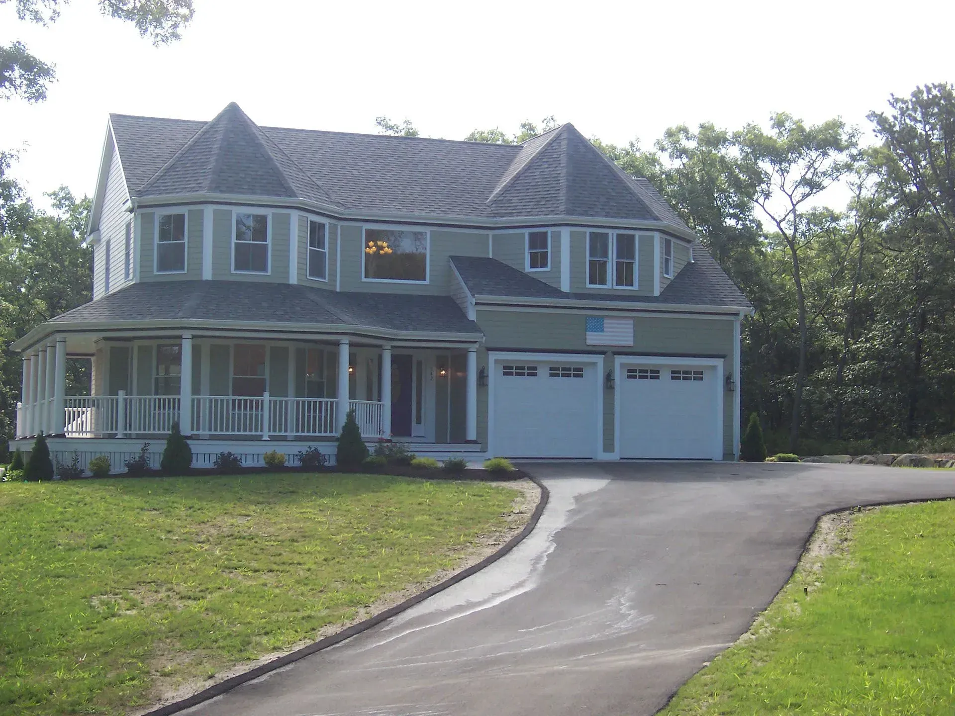 Two-story light green house with a curved porch, two-car garage, and asphalt driveway on a grassy lot.