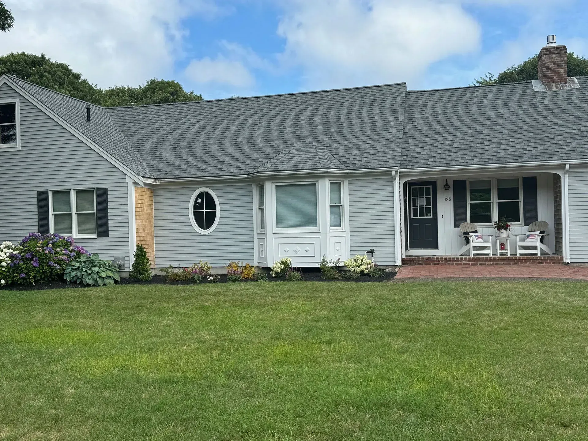 Gray-sided house with a dark roof and green lawn; white trim and an oval window are visible.