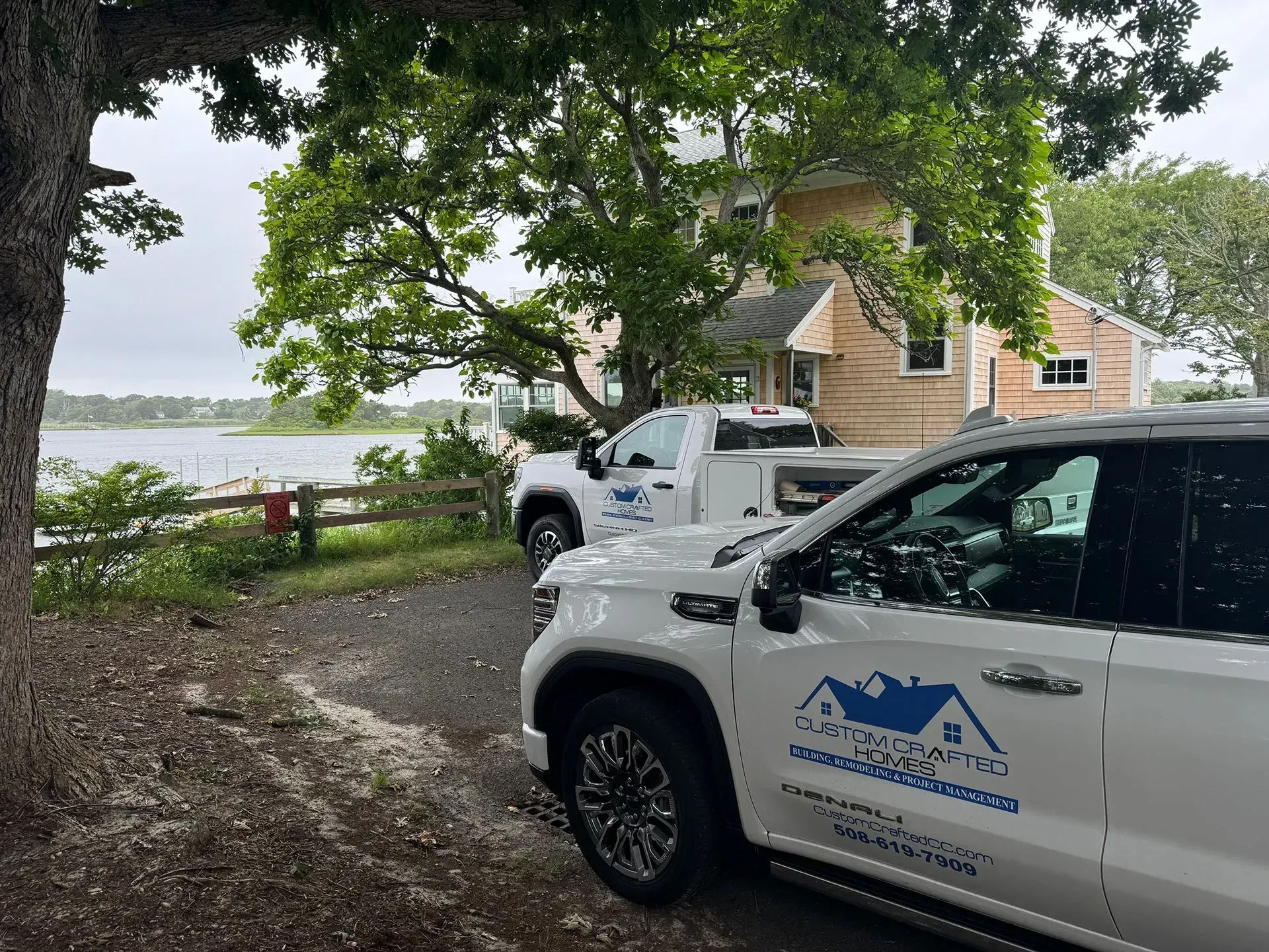 Two white trucks parked near a house, trees, and water. Trucks have a logo and business information on the side.