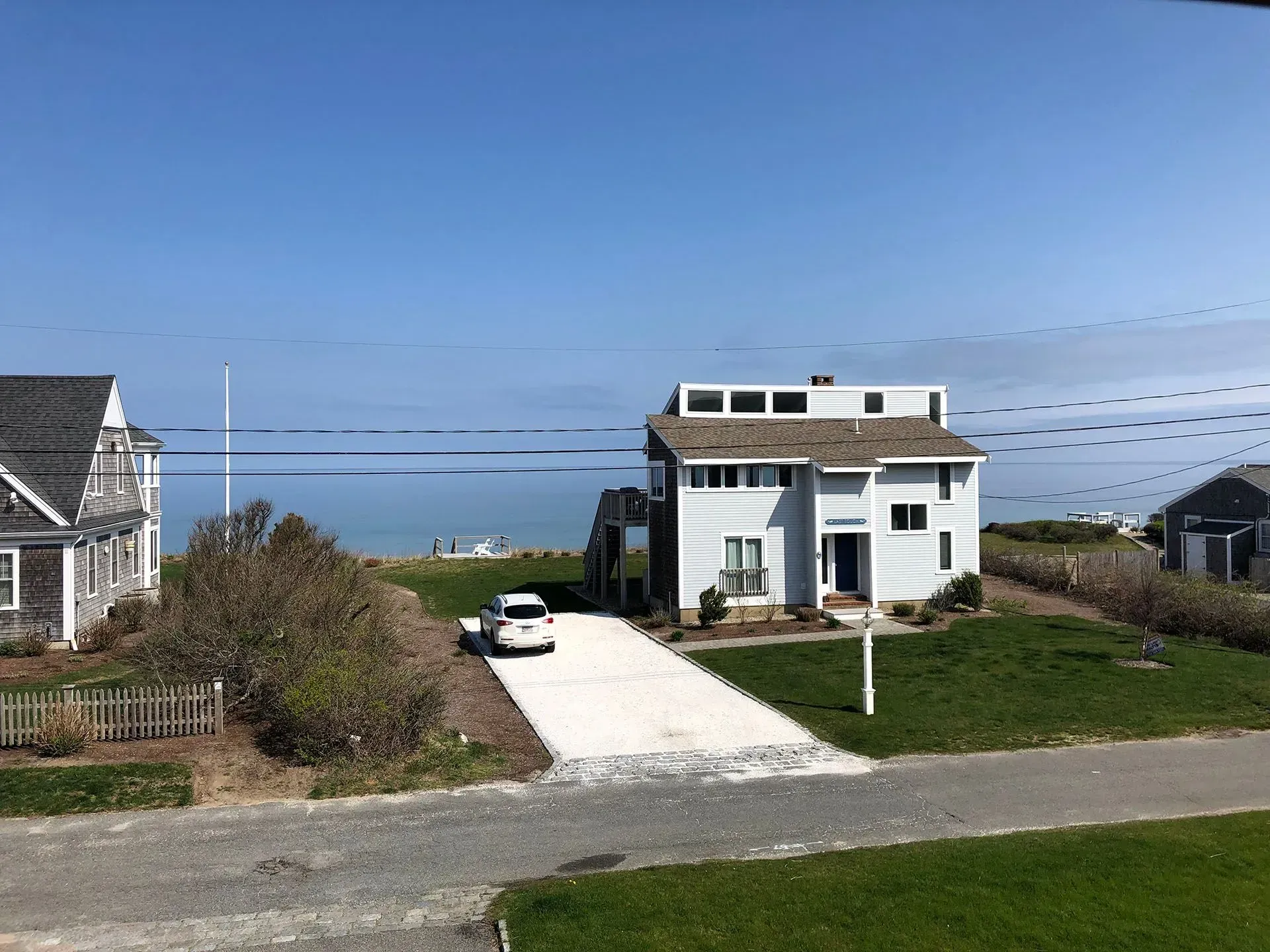 A light blue house with a gravel driveway and a white car sits on a grassy hill overlooking the ocean.