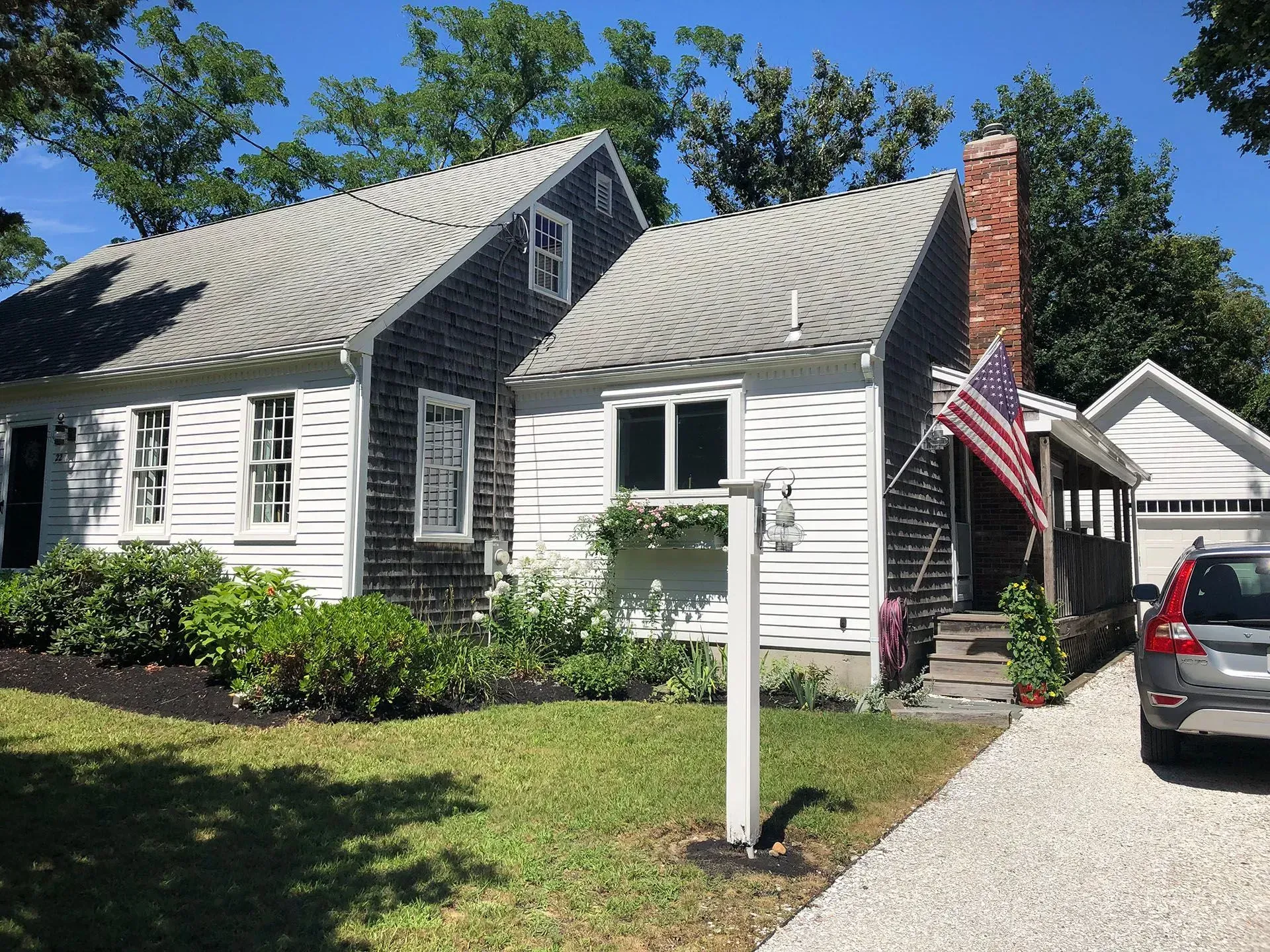 Cottage with white siding, dark shingles, American flag, brick chimney, gravel driveway, and a car.