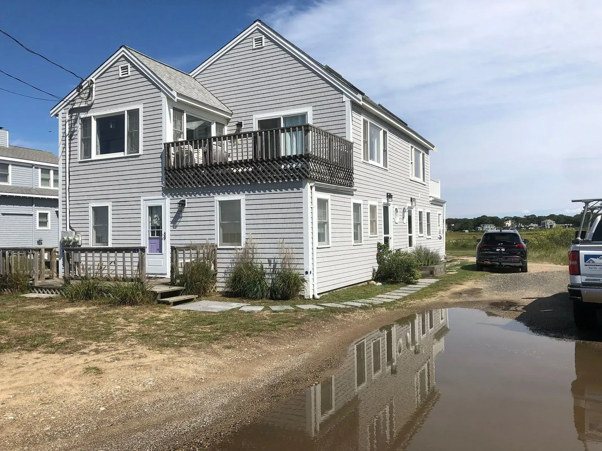 Two-story gray house with a small balcony, next to a flooded gravel driveway, parked vehicles.