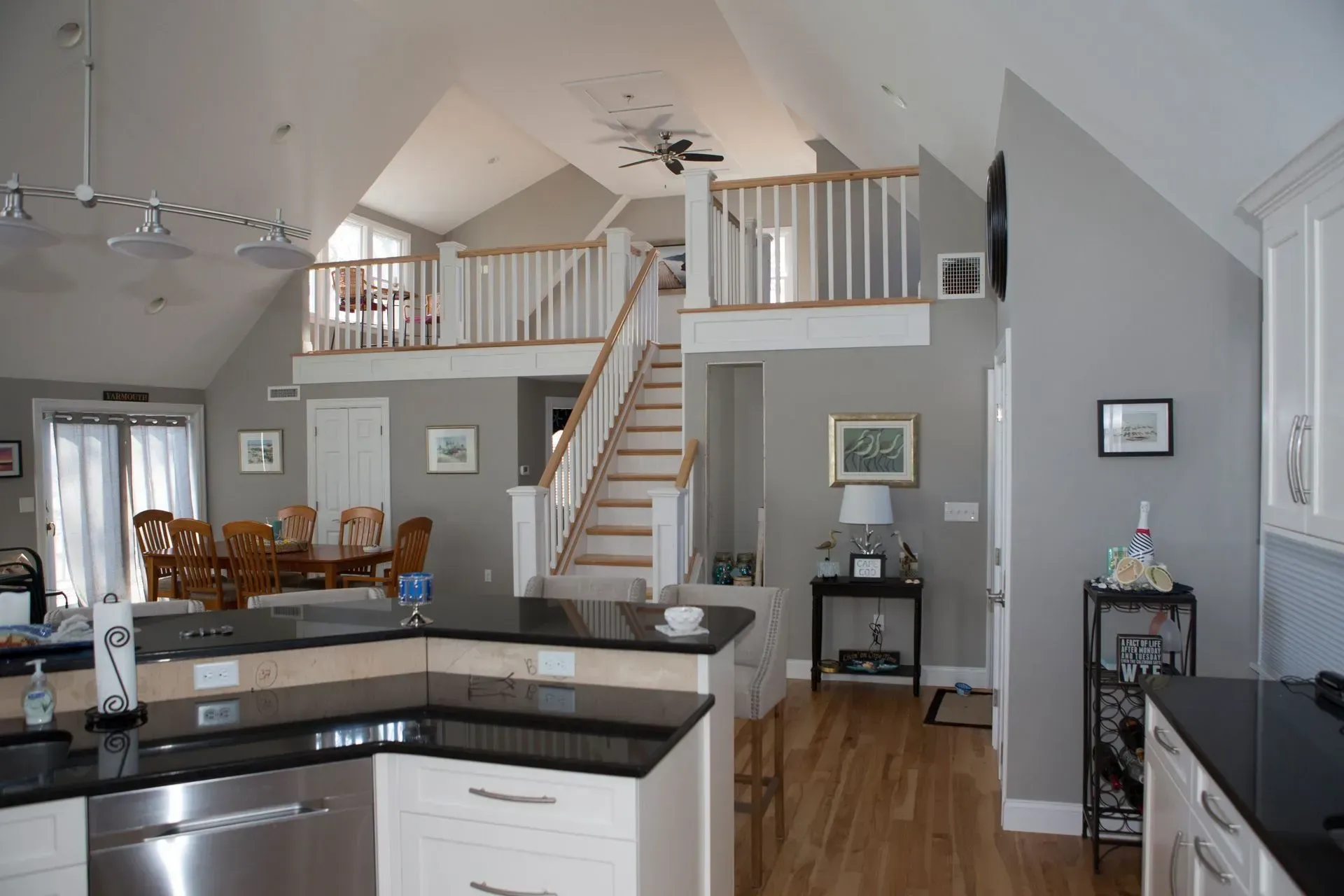 Kitchen interior with stairs to a loft; white cabinets, black countertops, wood floor, gray walls.