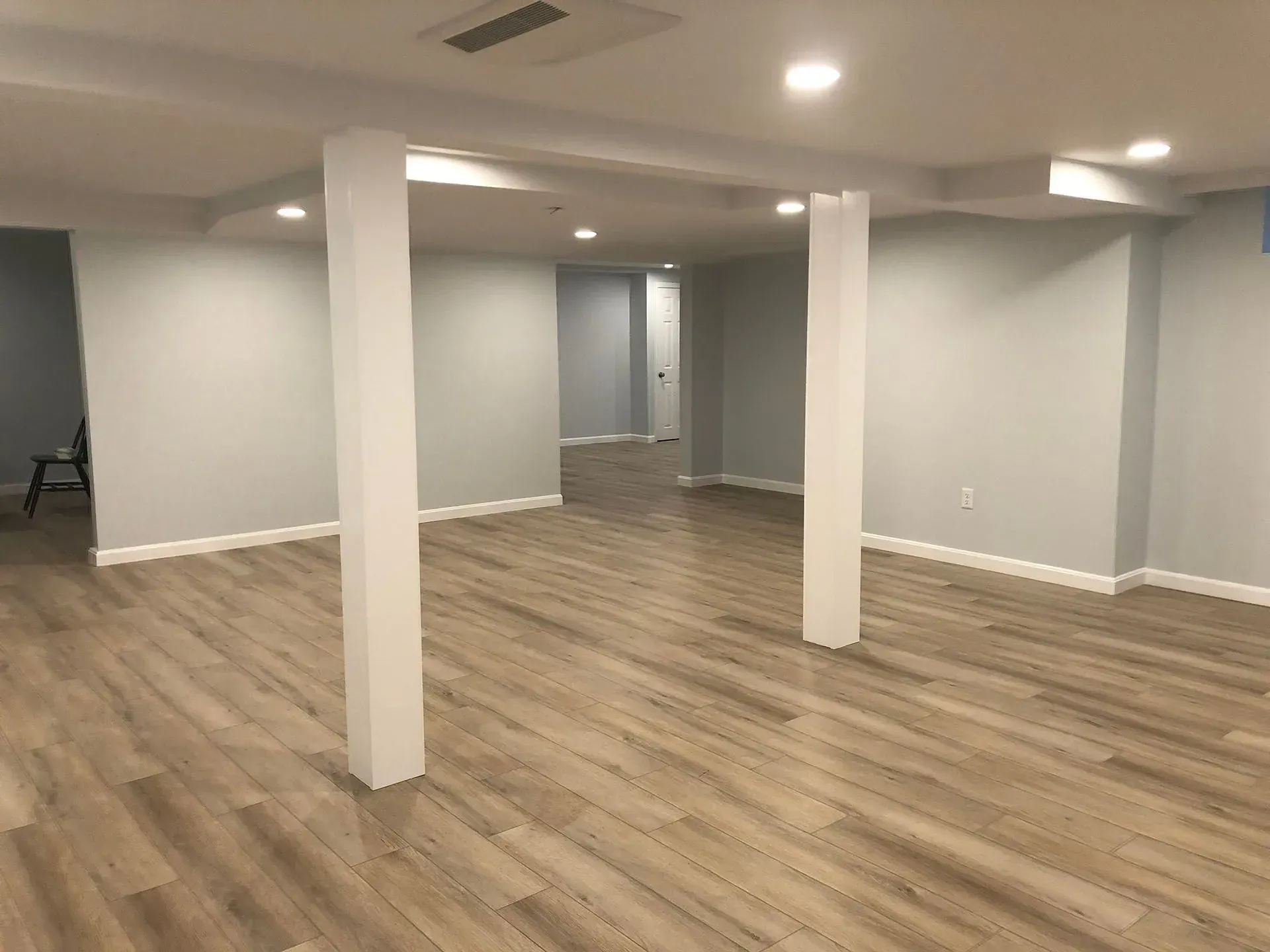 Empty, finished basement with light wood-look flooring, pale blue walls, white support columns, and recessed lighting.