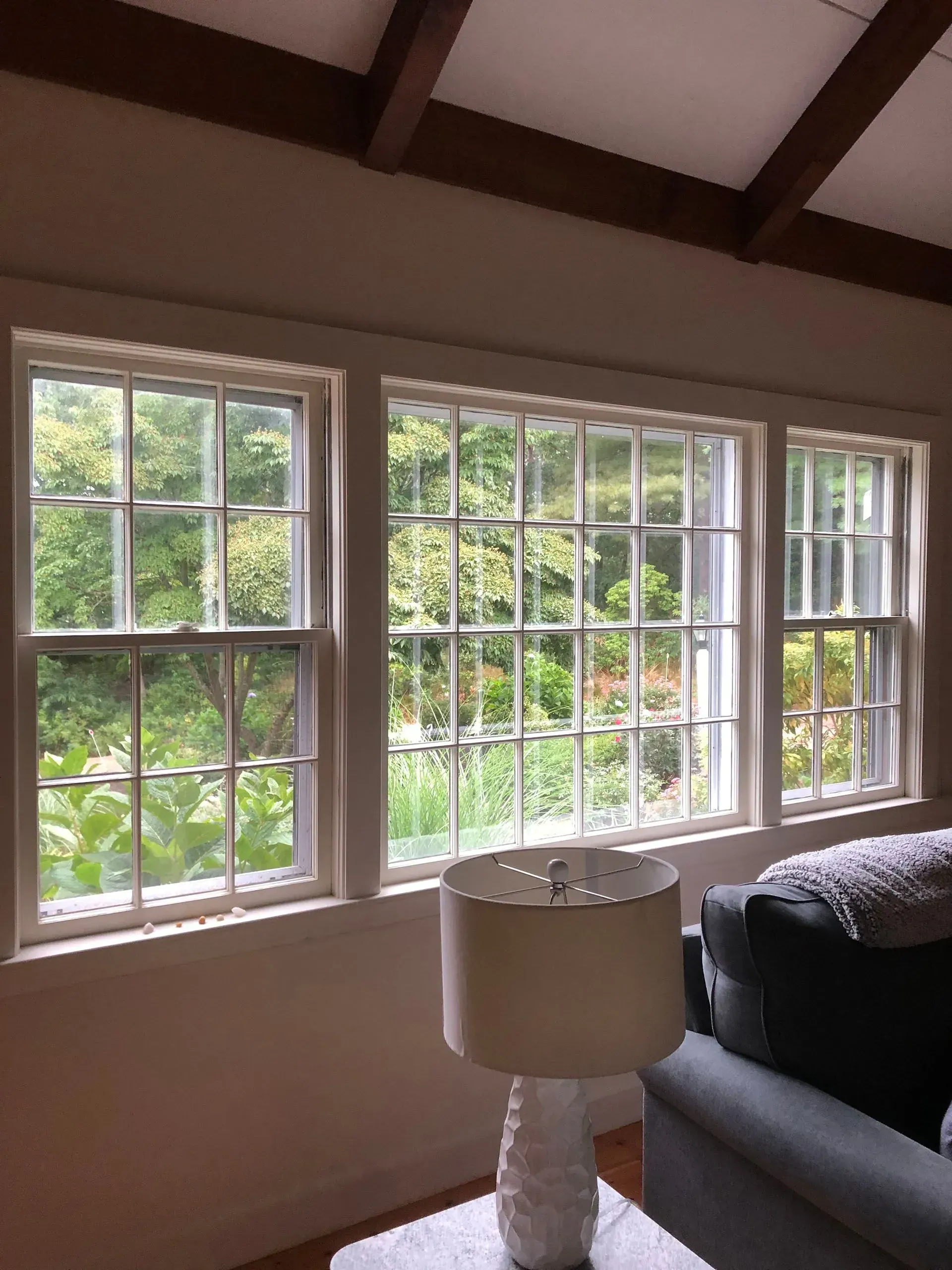 Three white-framed windows overlooking lush green foliage. A lamp is in front of a gray couch.