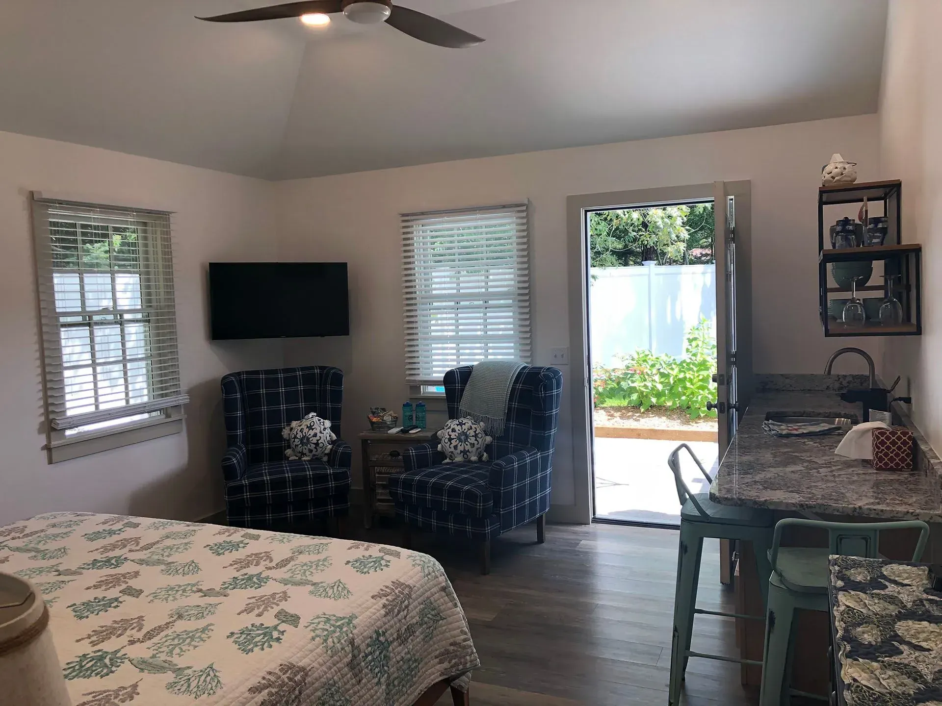 Cozy living space with bed, chairs, and kitchenette. Open doorway leads to greenery.