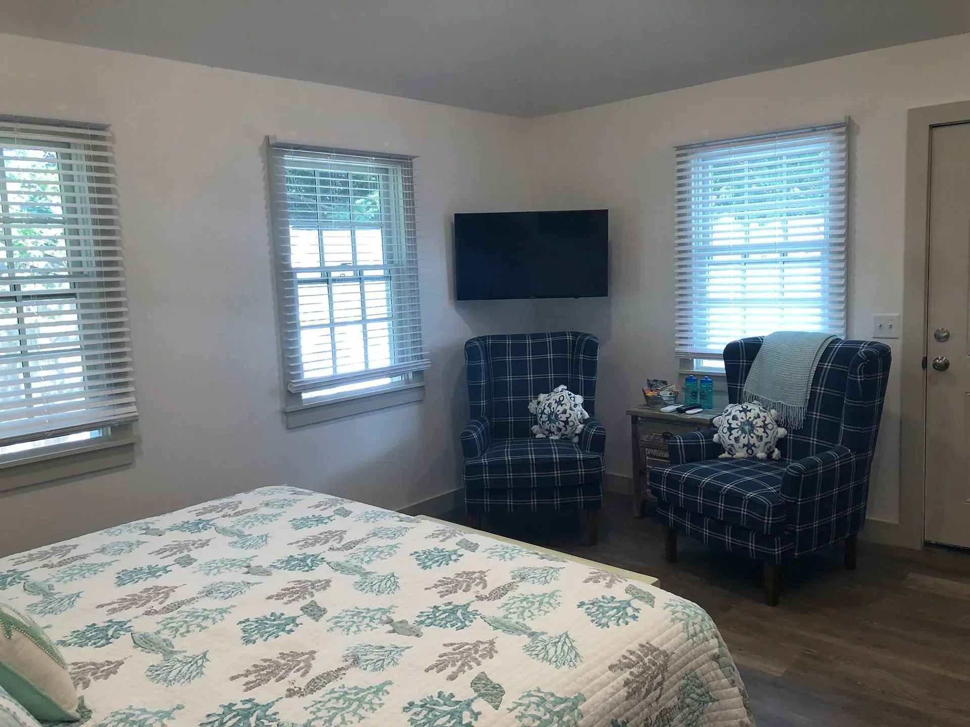 Bedroom with bed, patterned chairs, TV on wall, and two windows with blinds.