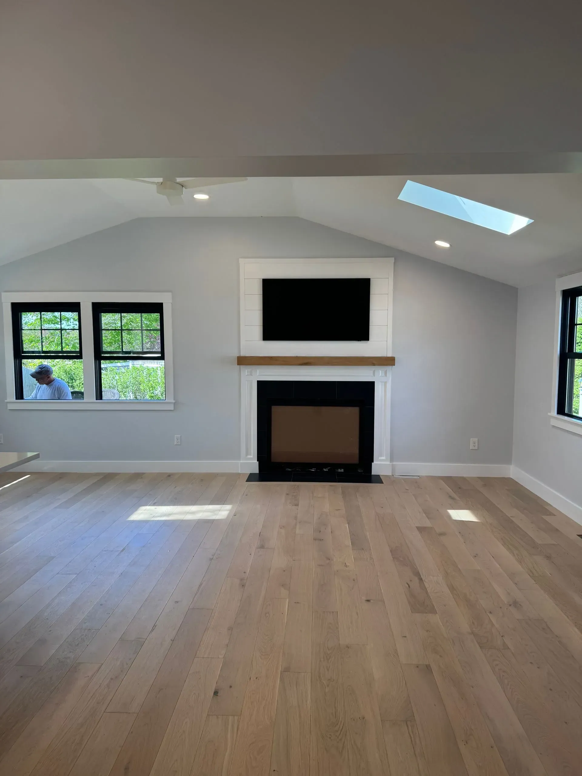 Living room with fireplace, TV, light wood floors, gray walls, and black-framed windows.