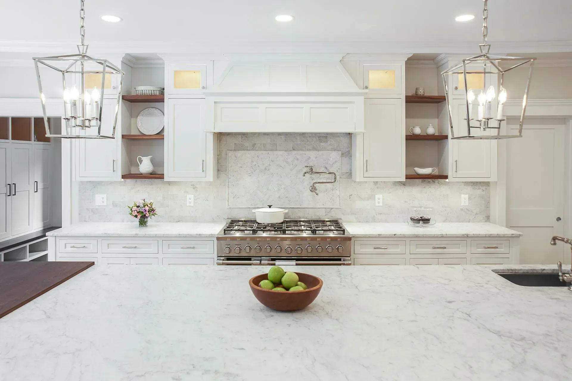 White kitchen with marble countertops, central island, gas stove, and hanging lights.