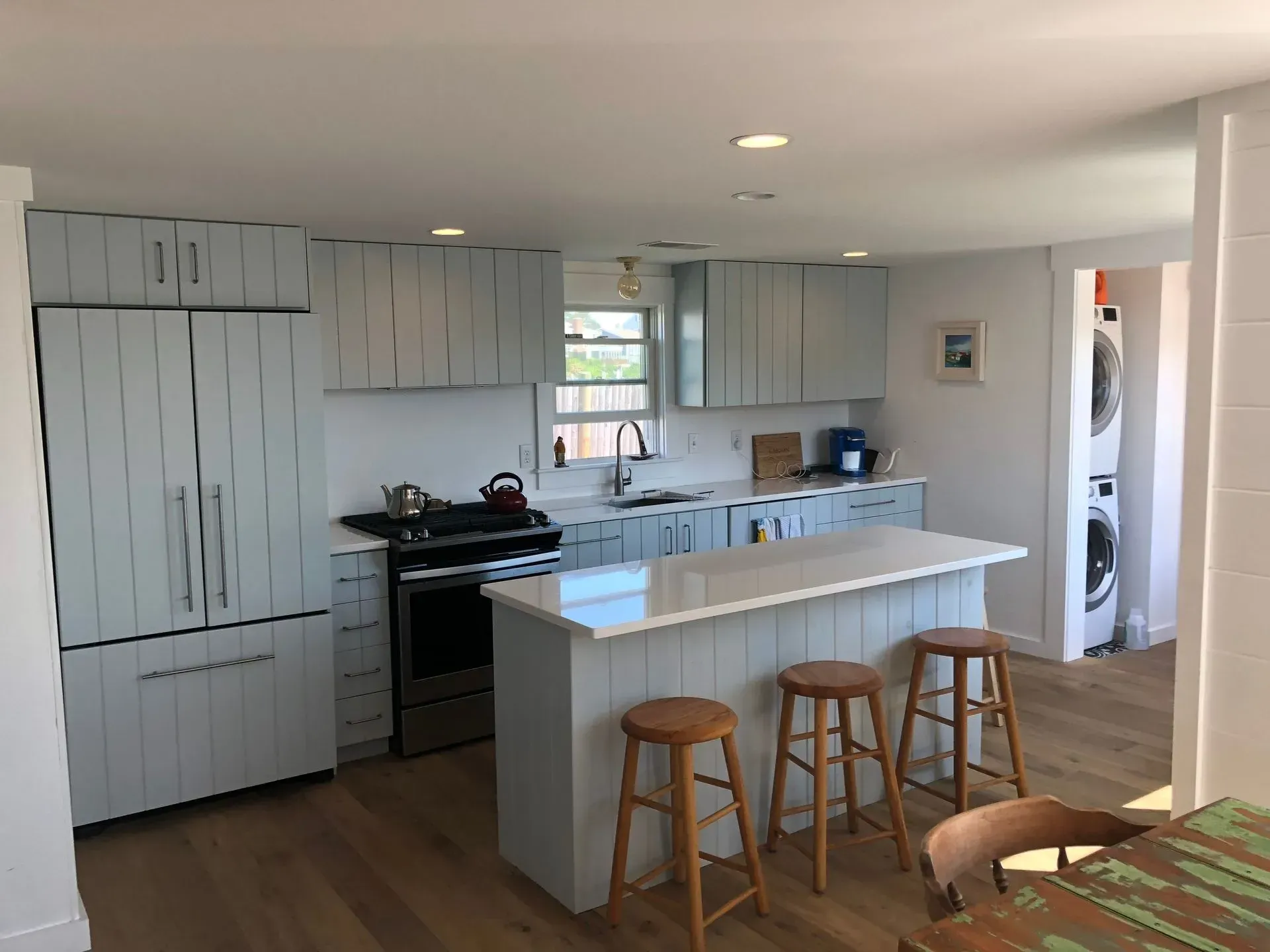 Light blue kitchen with island, stainless steel appliances, and wooden stools. Laundry room visible.