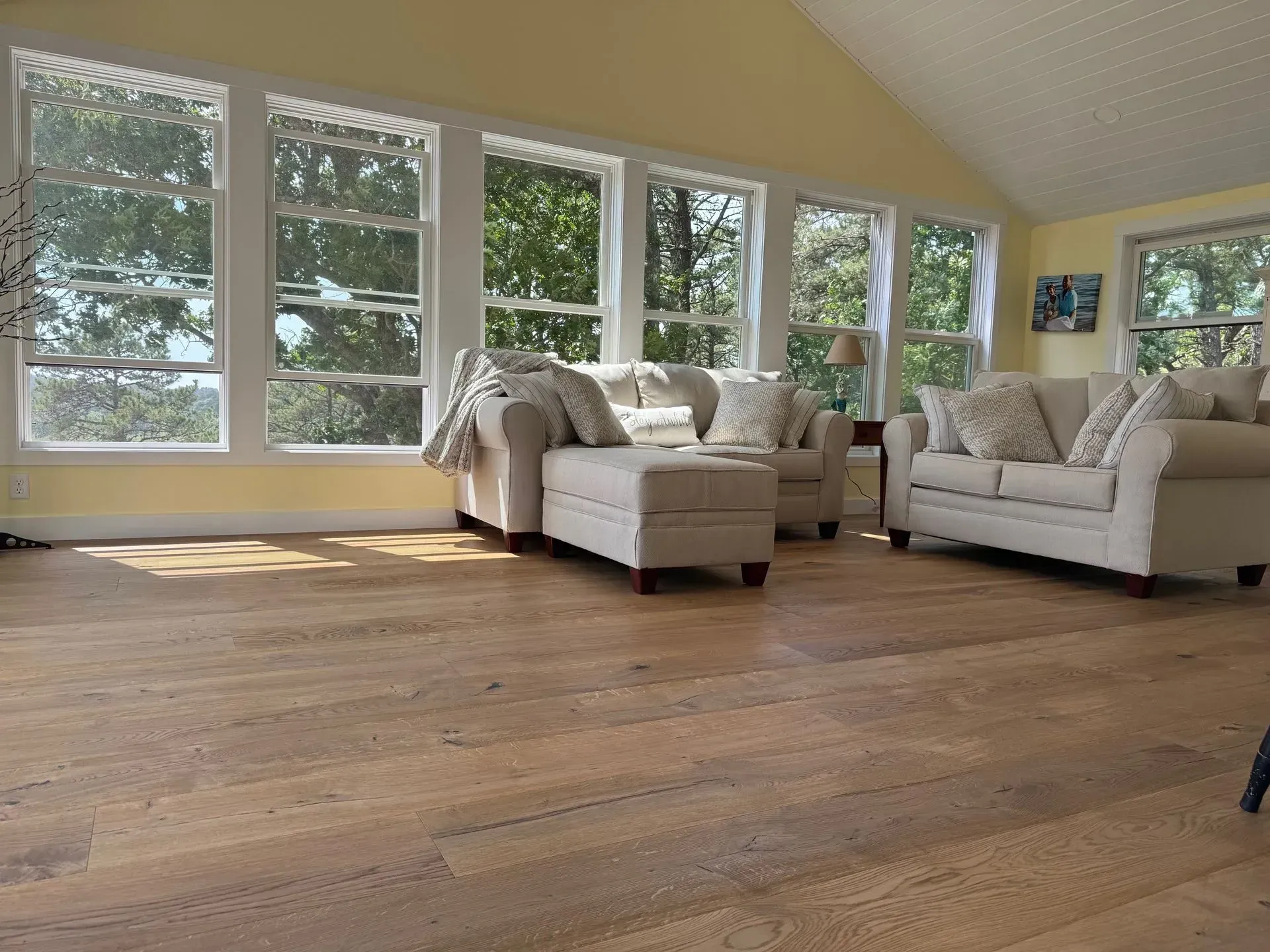 Living room with light wooden floors, white sofas, and large windows overlooking trees.