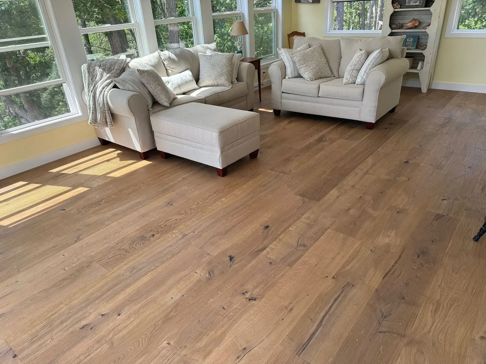 Sunlit living room with light wood floors, two couches with pillows, and an ottoman near windows.