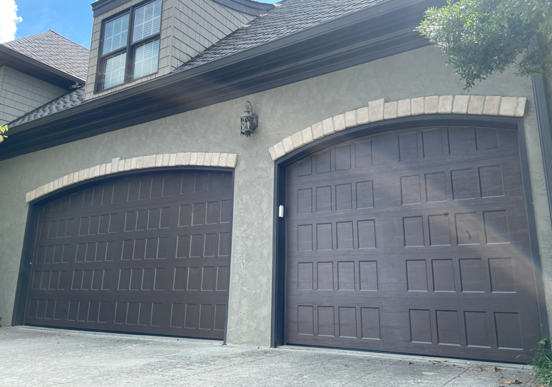 Two dark brown garage doors with decorative arched brick trim on a stucco house.