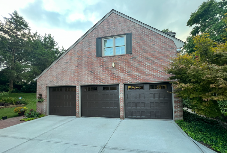 A detached brick garage with three dark brown doors and a single window above, set against a backdrop of trees.