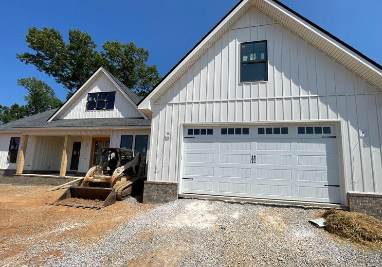 New construction of a white, board-and-batten farmhouse with a stone base, double garage, and a skid steer in the yard.