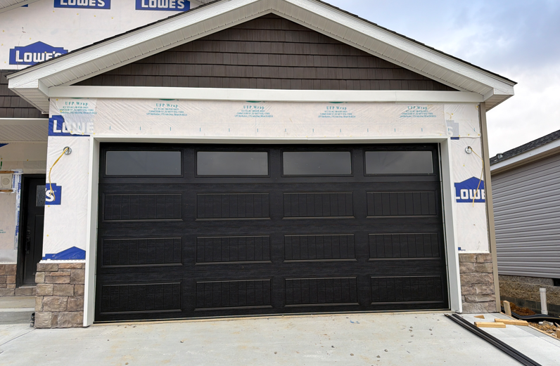 A black garage door with top windows installed on a house under construction with white siding and stone accents.
