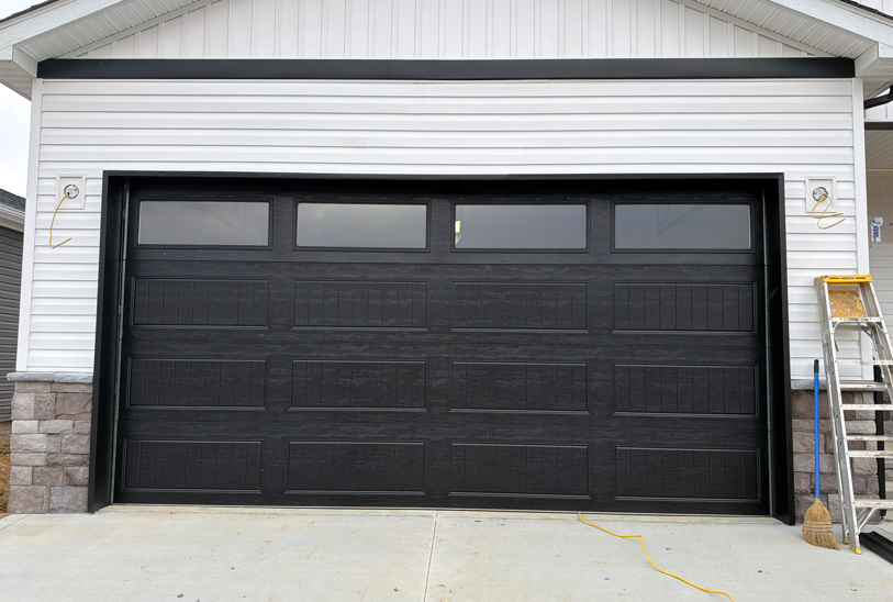 A black garage door with four top windows installed on a white house with stone accents and a ladder nearby.