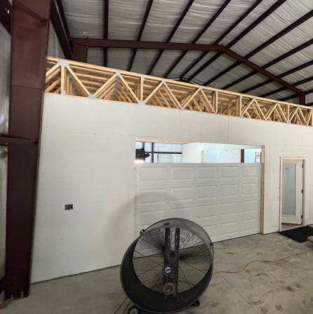 A white partition wall with a door and a garage door cutout, topped by an exposed wooden truss, in a large warehouse.
