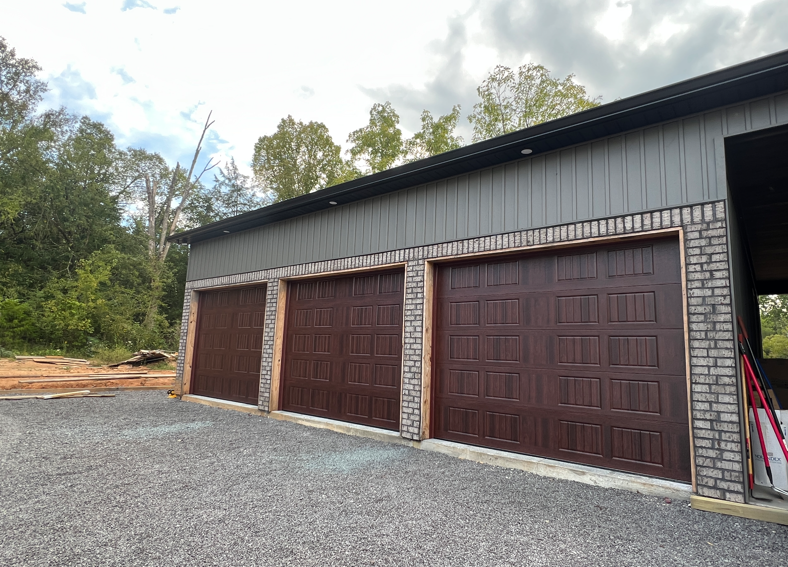 A modern detached three-car garage with brown doors, grey metal siding, and brick-style trim, set against a wooded area.