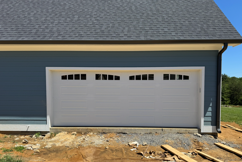 A white garage door with decorative top windows on a blue house under a clear sky, with dirt in the foreground.