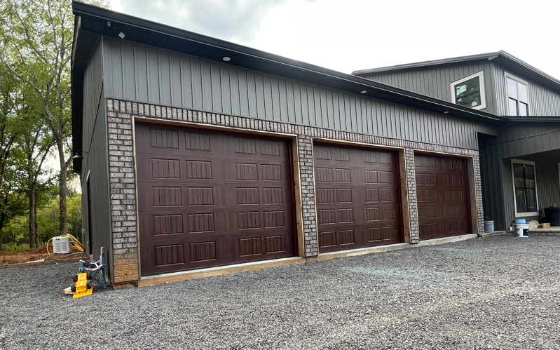 A large, modern garage featuring dark brown doors with brick accents and gray vertical siding on a gravel driveway.