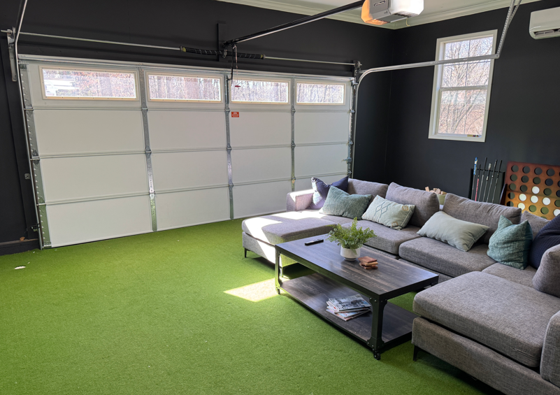 A garage converted into a lounge with dark walls, artificial grass flooring, a gray sectional sofa, and a coffee table.