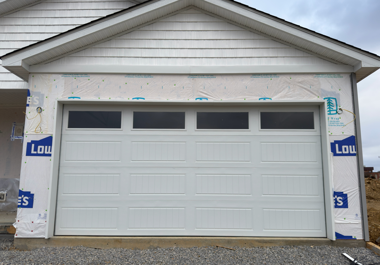 A white garage door with a row of top windows installed on an unfinished home under construction with blue house wrap.