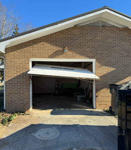 A white garage door hanging crookedly from one side on a brick building exterior.