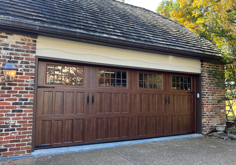 A dark wood, multi-panel garage door with small windows, set in a brick building with a dark shingled roof.