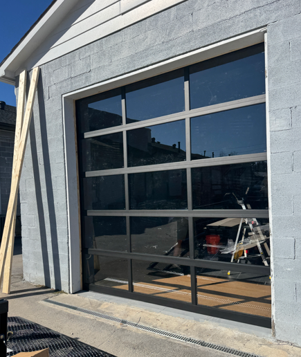 A modern, multi-paneled, dark-framed glass garage door installed in a grey cinder block building wall.