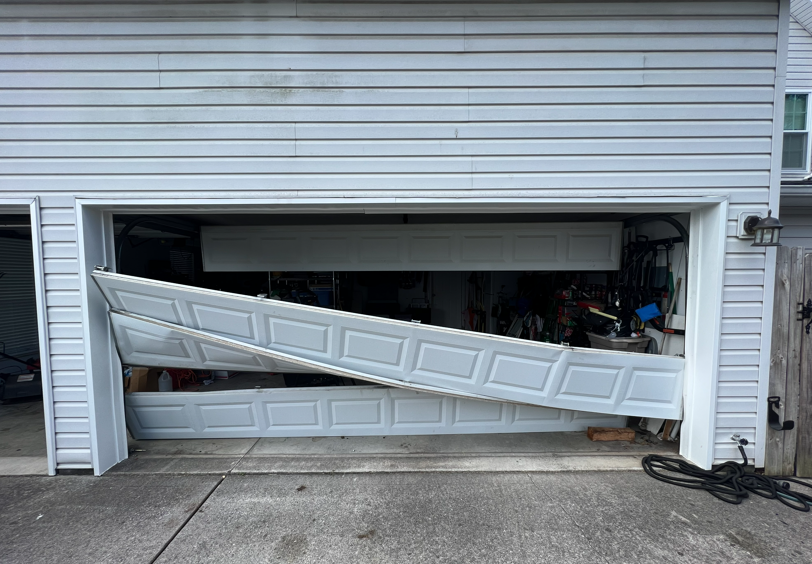 A white residential garage door has come off its tracks and is hanging at an angle across the open garage entrance.