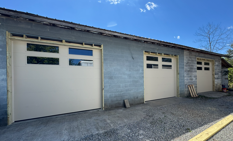 Three tan garage doors with rectangular windows installed on a grey concrete block building under a bright blue sky.