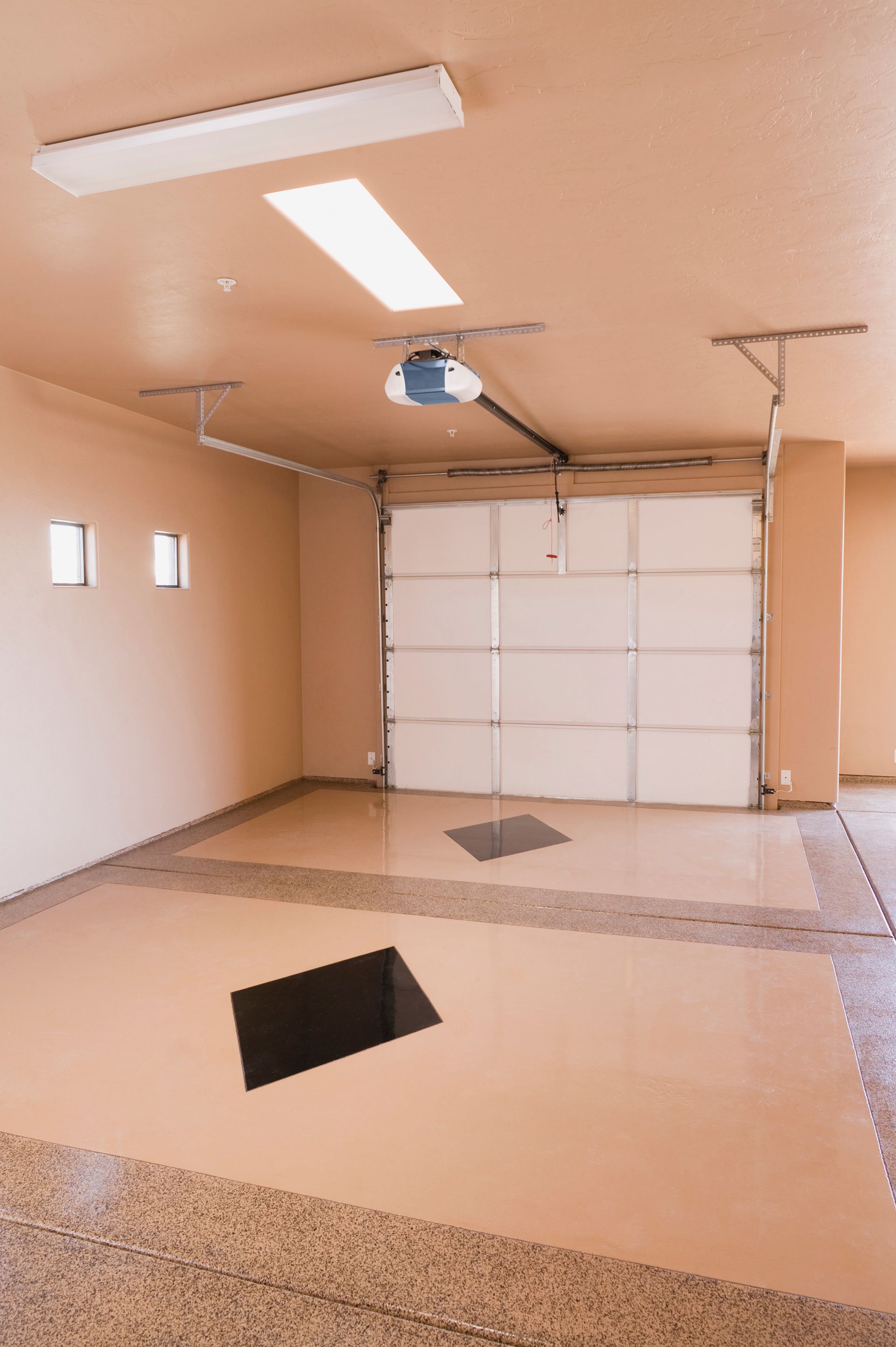 An empty garage with tan walls, white sectional door, overhead lighting, and tan epoxy flooring with black floor inlays.