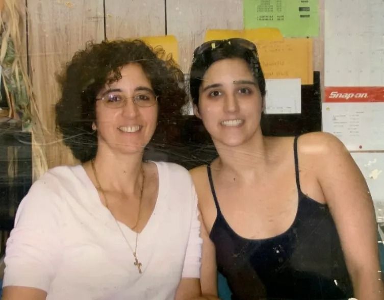 Two women smiling, posing together indoors. The woman on the left has curly hair and glasses; the other has short hair and is wearing a tank top.