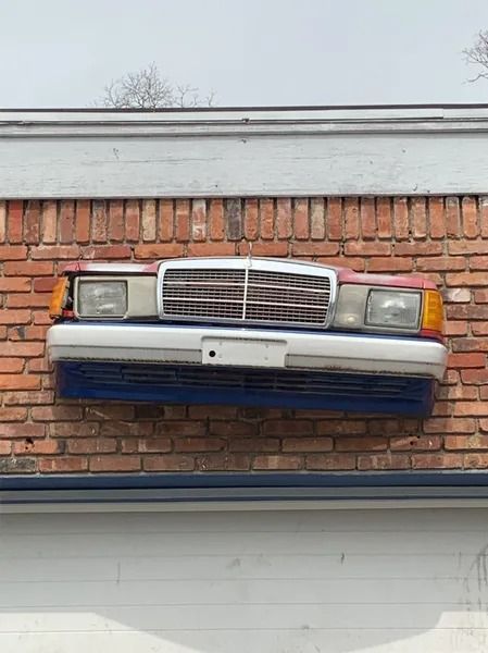 Car front end, featuring headlights, grill, and bumper, mounted on a red brick wall.