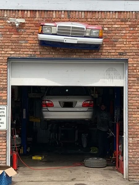 A car in a repair shop, lifted on a hoist. Above the garage door, a Mercedes-Benz front end is mounted on the brick wall.