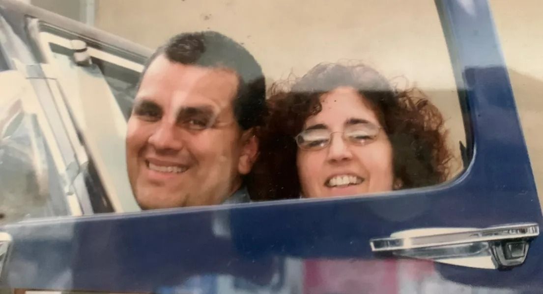 A man and woman smile from inside a blue vehicle. The man has dark hair, the woman glasses and curly hair.