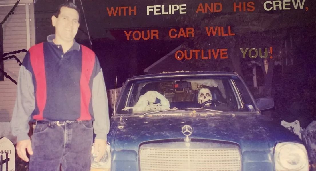 Man standing next to a blue car with two skeleton figures inside. Sign reads 