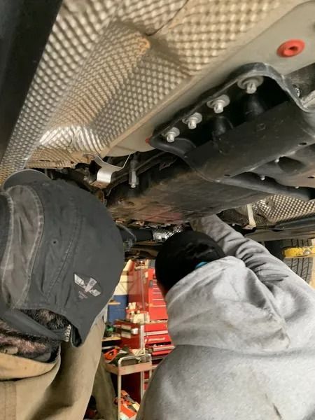 Two people working under a car. One points up at parts, likely performing repairs. Gray and silver car components are visible.